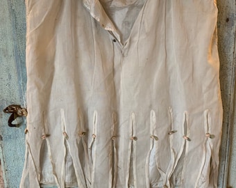 May include: A white sleeveless blouse with a button fastening and a delicate lace trim around the neckline. The blouse has a gathered detail at the bottom with small fabric loops and floral accents.