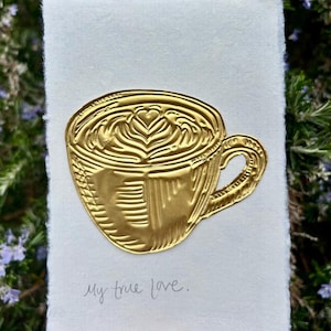 May include: A gold-coloured embossed artwork of a coffee cup with latte art on white paper. The text "My true love" is written below the cup. Signed "Katy Rose 2020".