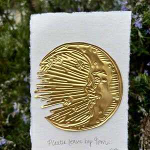 Puede incluir: Una luna creciente de metal dorado con una cara, estrellas y rayos de sol está fijada a un papel blanco texturizado. El texto "Please leave by 9pm" está escrito debajo. La firma del artista está en la esquina inferior derecha.