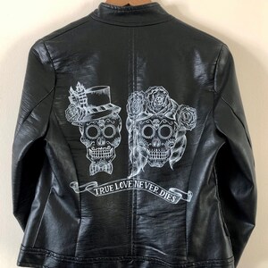 May include: Black leather jacket with a white design of two sugar skulls with floral accents and a banner that reads "TRUE LOVE NEVER DIES".
