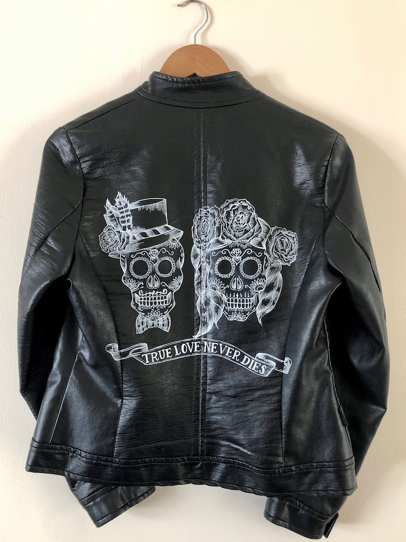 May include: Black leather jacket with a white design of two sugar skulls with floral accents and a banner that reads "TRUE LOVE NEVER DIES".