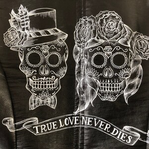 May include: Black leather jacket with a white design of two sugar skulls with floral accents. The text "TRUE LOVE NEVER DIES" is written in a banner below the skulls.