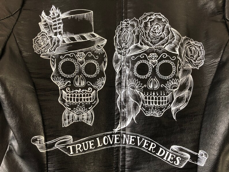 May include: Black leather jacket with a white design of two sugar skulls with floral accents. The text "TRUE LOVE NEVER DIES" is written in a banner below the skulls.