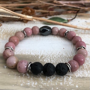 May include: A pink and black beaded bracelet with silver accents. The bracelet features round pink beads, two black lava beads, and a single silver bead with a black oval-shaped stone.