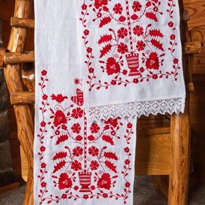 May include: Two white linen table runners with red embroidered floral designs and white lace trim. The runners are draped over a wooden chair.