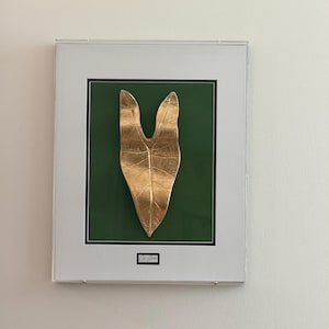Pode incluir: Uma peça de arte de parede emoldurada com uma escultura de folha em tom dourado metálico centrada num fundo verde profundo. A obra está inserida numa moldura branca com um passe-partout branco e inclui uma pequena placa com uma assinatura.