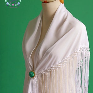 May include: White shawl with a crocheted border and fringe. The shawl is decorated with a green flower brooch.