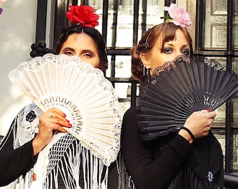 May include: Two women wearing black dresses and lace shawls stand behind a black metal railing. One woman holds a white lace fan in front of her face, while the other holds a black lace fan in front of her face. Both women are wearing flowers in their hair.