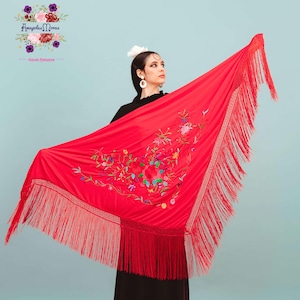 Puede incluir: Un chal rojo vibrante con intrincados bordados florales en varios colores. El chal presenta largas y a juego flecos en los bordes. La imagen también muestra el logotipo "Amapolas Moras" y "tienda flamenca".