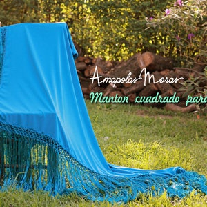 Op de afbeelding: Een felblauwe sjaal met franjes, gedrapeerd over een houten stoel. De sjaal is gelabeld met "Amapolas Moran Manton cuadrado para Baile".