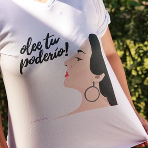 May include: White t-shirt with black text that says "olee tu poderio!" and a black and white illustration of a woman with red lips and a large hoop earring.