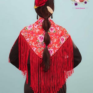 Puede incluir: Un chal flamenco rojo con estampado floral y flecos largos. El chal se lleva sobre una prenda negra. La imagen también muestra el cabello peinado en una trenza, adornado con una flor roja y accesorios. El texto "Amapolas Moras" y "tienda flamenca" es visible.