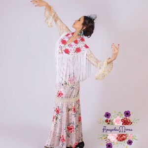 May include: A woman wearing a white lace top with floral embroidery and a white fringe, a floral print skirt, and black shoes. She is posing with her arms outstretched. The image includes the text "Amapolas Moras tienda Flamenca".