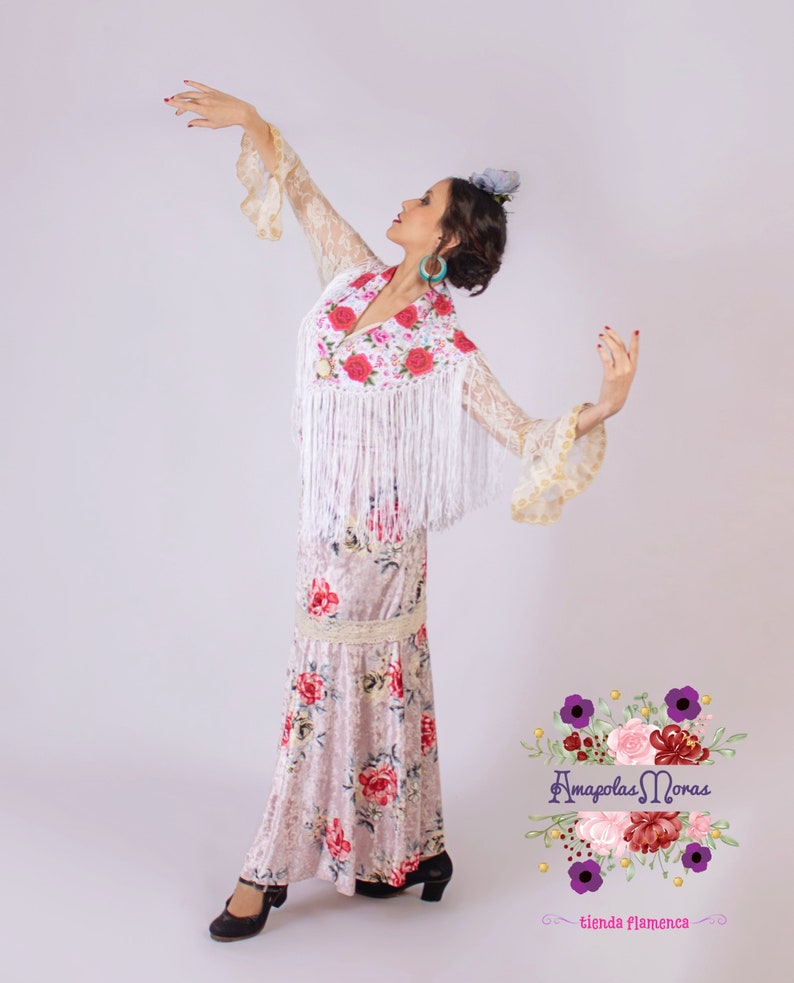 May include: A woman wearing a white lace top with floral embroidery and a white fringe, a floral print skirt, and black shoes. She is posing with her arms outstretched. The image includes the text "Amapolas Moras tienda Flamenca".