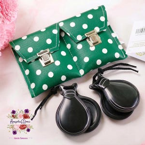Puede incluir: Un bolso de mano verde con lunares blancos, un cierre plateado y dos castañuelas negras con cordones negros. El bolso y las castañuelas están sobre una superficie rosa. El bolso tiene un cierre de solapa.