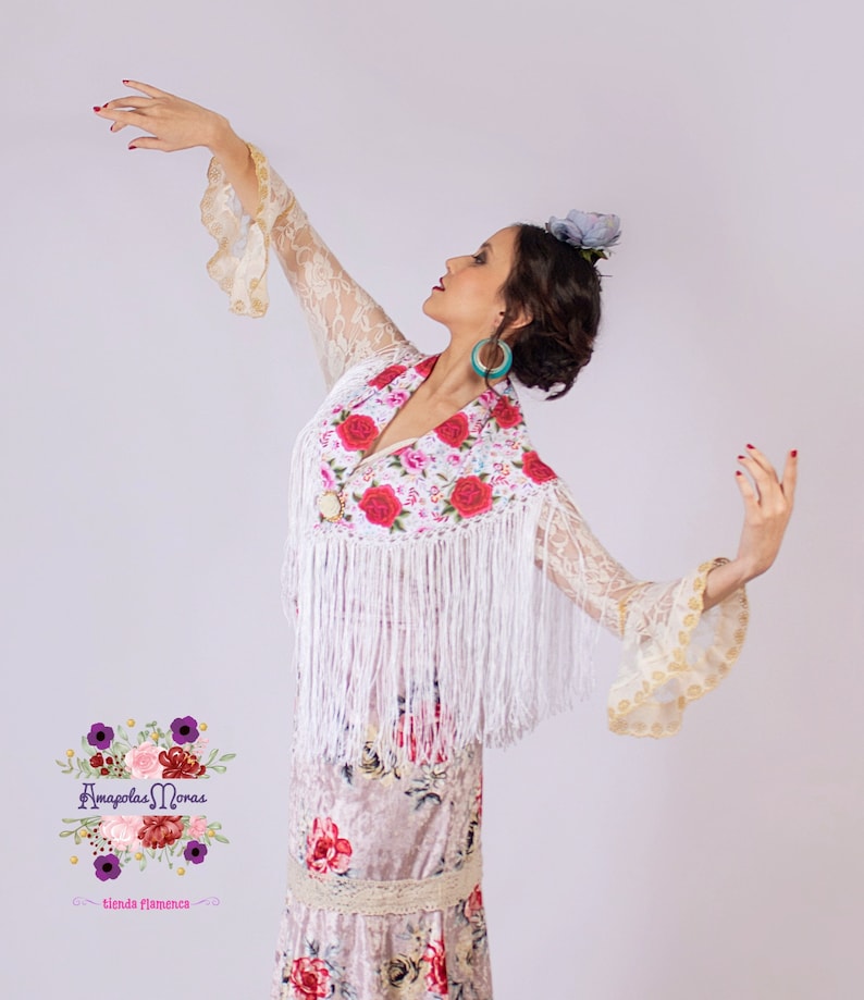 May include: A woman wearing a white lace dress with a floral pattern and a white shawl with fringe. She is wearing a flower in her hair and has her arms outstretched. The text "Amapola Moras - tienda flamenca" is visible in the lower left corner of the image.