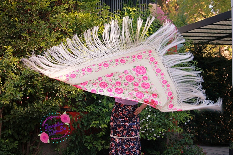 Op de afbeelding: Een cr&egrave;mekleurige sjaal met een bloemmotief van roze rozen en groene bladeren, omlijst door een witte franje. De sjaal wordt omhoog gehouden, de franje wappert in de wind. De achtergrond is een weelderige groene tuin.