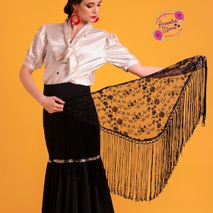 Op de afbeelding: Een zwarte kant shawl met bloemenborduurwerk en franjes. De shawl is over de arm van een persoon gelegd die een witte polka dot blouse en een zwarte rok draagt.