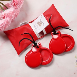 Puede incluir: Castañuelas rojas con cuerdas negras y una funda de cuero roja. La funda tiene una etiqueta blanca con texto y un logotipo. Las castañuelas son un instrumento musical tradicional español.