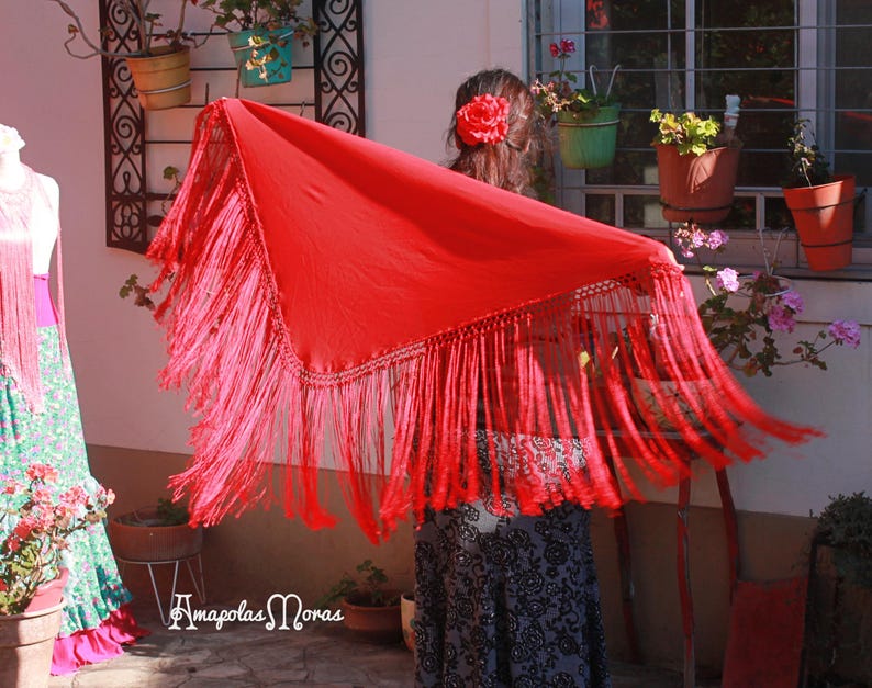 May include: A red shawl with fringe, worn by a person with a flower in their hair. The shawl is draped over the person's shoulders and the fringe is flowing in the wind.