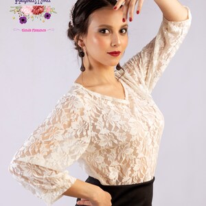 Puede incluir: Una blusa blanca de encaje con mangas 3/4. La blusa tiene un escote en V y un delicado patrón floral. La modelo lleva una falda negra y tiene el pelo recogido hacia atrás.