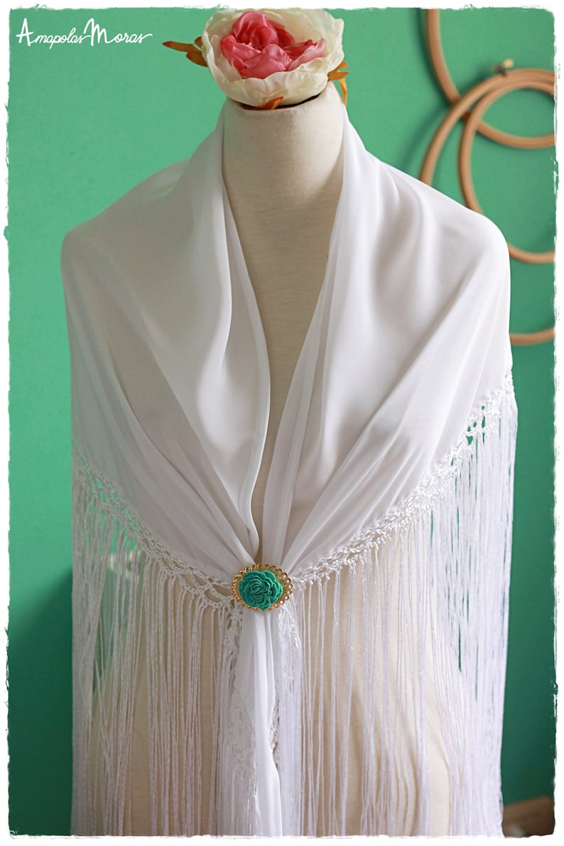May include: White shawl with fringe and a turquoise flower brooch. The shawl is draped over a white mannequin.
