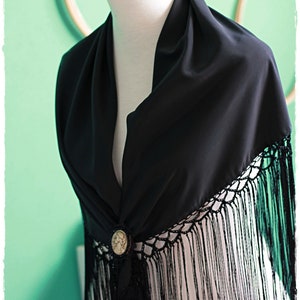 May include: Black shawl with a crocheted fringe and a decorative button. The shawl is draped over a mannequin with a pink rose on top.