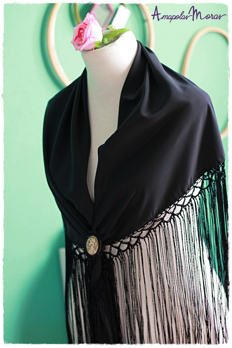 May include: Black shawl with a crocheted fringe and a decorative button. The shawl is draped over a mannequin with a pink rose on top.