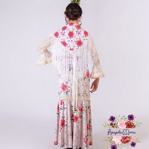 May include: A white lace shawl with floral embroidery and white fringe, worn over a pink floral dress. The dress has a ruffled hem and a lace trim. The model is wearing a flower in her hair.