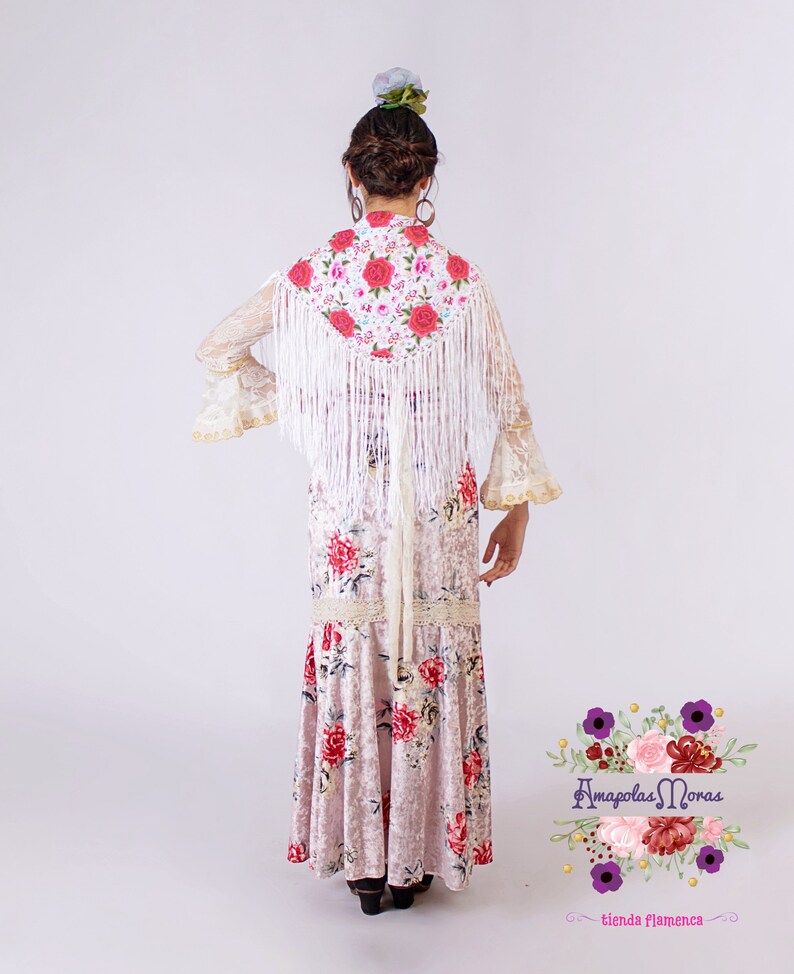 May include: A white lace shawl with floral embroidery and white fringe, worn over a pink floral dress. The dress has a ruffled hem and a lace trim. The model is wearing a flower in her hair.