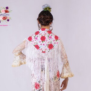 May include: White shawl with a floral pattern and fringe. The shawl is embroidered with red and pink roses. The shawl is worn over a white lace dress.