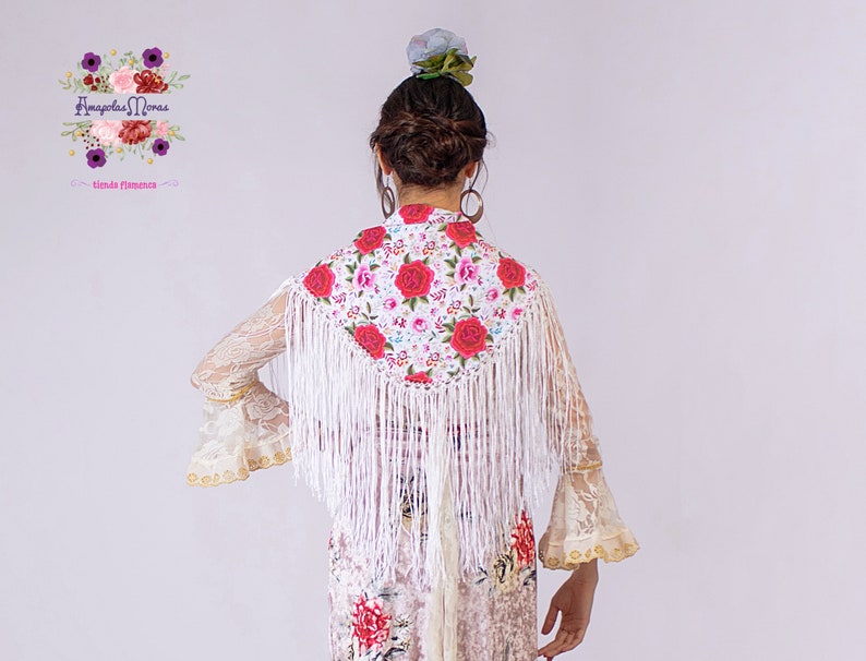 May include: White shawl with a floral pattern and fringe. The shawl is embroidered with red and pink roses. The shawl is worn over a white lace dress.