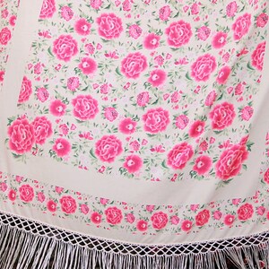 Op de afbeelding: Een cr&egrave;mekleurige sjaal met een levendig bloemmotief van grote roze pioenrozen en kleinere roze bloemen. De sjaal heeft een witte franjebies en een decoratieve rand van roze bloemen. Het ontwerp is vrouwelijk en elegant.