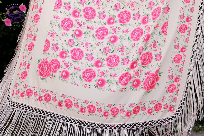 Op de afbeelding: Een cr&egrave;mekleurige sjaal met een levendig bloemmotief van grote roze pioenrozen en kleinere roze bloemen. De sjaal heeft een witte franjebies en een decoratieve rand van roze bloemen. Het ontwerp is vrouwelijk en elegant.