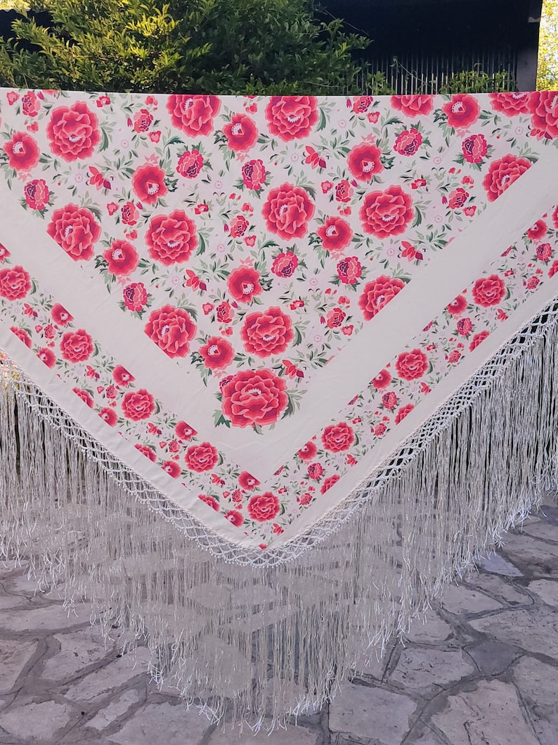 Op de afbeelding: Een cr&egrave;mekleurige sjaal met een bloemmotief van rode rozen en groene bladeren. De sjaal heeft een witte rand en lange, witte franjes. Het ontwerp doet denken aan traditionele Spaanse sjaals. De stof is licht.