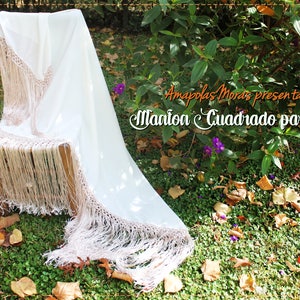Op de afbeelding: Een witte sjaal met een franje rand, gedrapeerd over een houten stoel. De sjaal is gemaakt van een lichtgewicht stof en heeft een delicaat kantpatroon. De tekst "Amapola Moras presenta su Manion Cuadrado para Baile" is zichtbaar op de achtergrond.