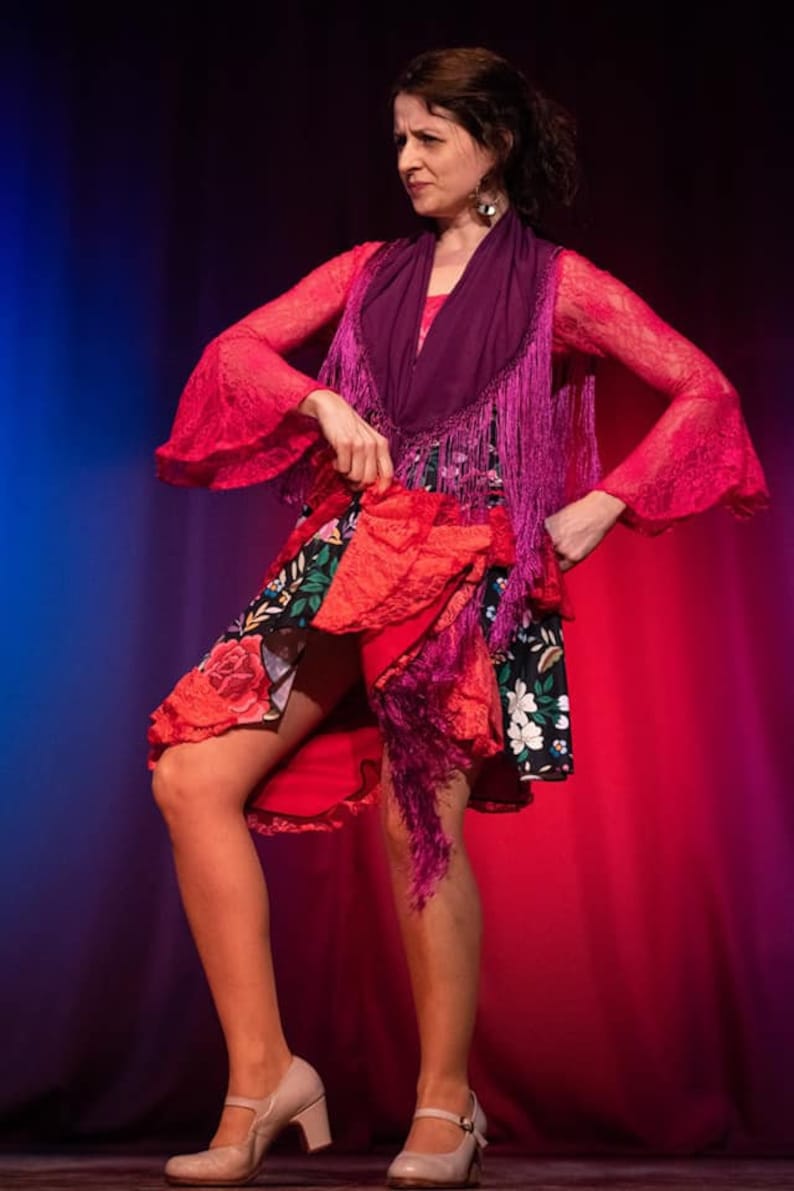 May include: A woman in a pink and purple flamenco dress with a floral print skirt and fringe accents performs on stage.