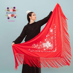 Puede incluir: Un chal rojo vibrante con bordados florales blancos y largas borlas. El chal se sostiene en alto, mostrando el intrincado diseño. El fondo es de un azul claro sólido.