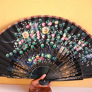 Puede incluir: Un abanico de mano negro con diseños florales dorados y verdes pintados en él. El abanico está cerrado y está siendo sostenido por una mano.