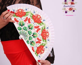 May include: White hand fan with a red, green, and blue floral design. The fan is closed and held by a person's hand. The text "Amapola Moras" and "tienda flamenca" is visible in the background.
