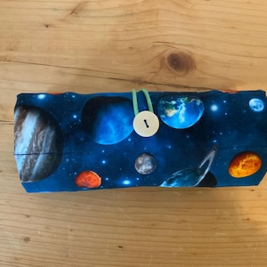 Könnte beinhalten: Eine zylindrische Stoffrolle mit einem dunkelblauen Weltraumdruck, der verschiedene Planeten, Sterne und Galaxien zeigt. Die Rolle wird mit einer hellgrünen elastischen Kordel gesichert, die um einen einzelnen weißen, runden Knopf geschlungen ist.