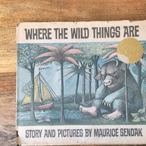 May include: A vintage hardcover book titled "Where the Wild Things Are" with illustrations of a blue monster and a sailboat. The book cover is blue, green, and yellow with a gold sticker on the front.