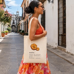 Puede incluir: Una bolsa de tela color crema con una ilustraci&oacute;n gr&aacute;fica de dos rodajas de pl&aacute;tano frito y el texto: tost&oacute;n, Rodaja de pl&aacute;tano verde, machacado y frito. Sin&oacute;nimo: patac&oacute;n. La bolsa tiene dos asas para el hombro y una forma rectangular.