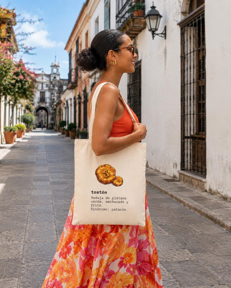 Puede incluir: Una bolsa de tela color crema con una ilustraci&oacute;n gr&aacute;fica de dos rodajas de pl&aacute;tano frito y el texto: tost&oacute;n, Rodaja de pl&aacute;tano verde, machacado y frito. Sin&oacute;nimo: patac&oacute;n. La bolsa tiene dos asas para el hombro y una forma rectangular.