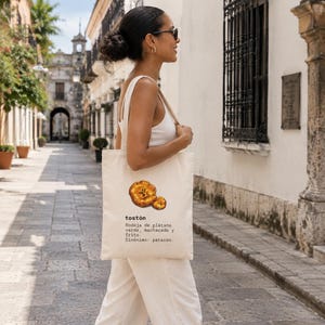 Puede incluir: Una bolsa de tela color crema con una ilustraci&oacute;n gr&aacute;fica de dos rodajas de pl&aacute;tano frito. El texto dice: tost&oacute;n, Rodaja de pl&aacute;tano verde, machacado y frito, Sin&oacute;nimo: patac&oacute;n. La bolsa tiene dos asas para el hombro y una forma rectangular.
