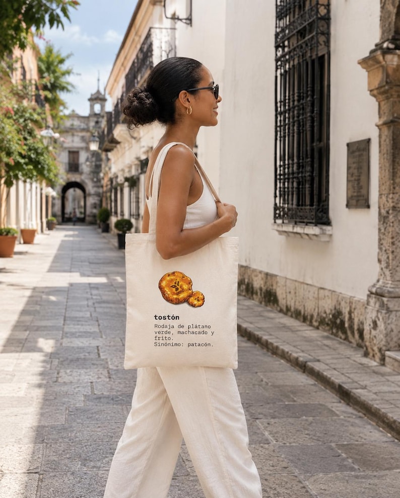 Puede incluir: Una bolsa de tela color crema con una ilustraci&oacute;n gr&aacute;fica de dos rodajas de pl&aacute;tano frito. El texto dice: tost&oacute;n, Rodaja de pl&aacute;tano verde, machacado y frito, Sin&oacute;nimo: patac&oacute;n. La bolsa tiene dos asas para el hombro y una forma rectangular.