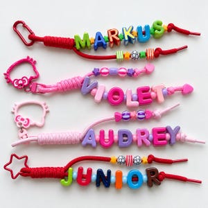 Può includere: Quattro portachiavi con perline colorate che riportano nomi composti da lettere in plastica. Il portachiavi in alto è rosso con il nome MARKUS, il secondo è rosa con VIOLET, il terzo è rosa chiaro con AUDREY e quello in basso è rosso con JUNIOR.