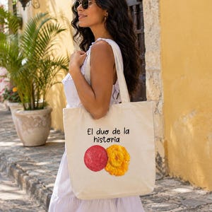 Puede incluir: Una bolsa de tela color crema con el texto negro "El duo de la historia" sobre una ilustraci&oacute;n gr&aacute;fica de una rodaja de salami roja y una rodaja de pl&aacute;tano frito amarillo dorado, con dos asas para el hombro y una forma rectangular.