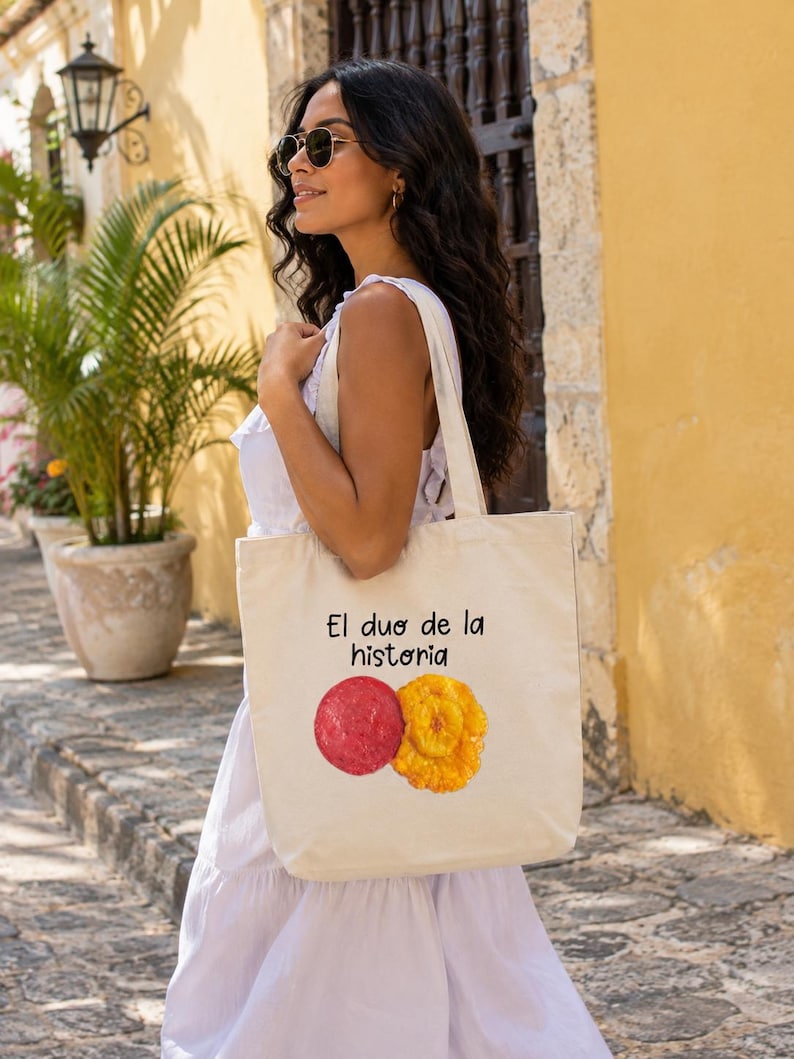 Puede incluir: Una bolsa de tela color crema con el texto negro "El duo de la historia" sobre una ilustraci&oacute;n gr&aacute;fica de una rodaja de salami roja y una rodaja de pl&aacute;tano frito amarillo dorado, con dos asas para el hombro y una forma rectangular.