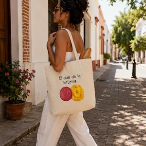 Puede incluir: Una bolsa de tela beige con el texto negro El duo de la historia sobre un gr&aacute;fico de una rodaja de salami roja circular y un grupo amarillo de chips de pl&aacute;tano frito. La bolsa cuenta con dos asas para llevar al hombro.
